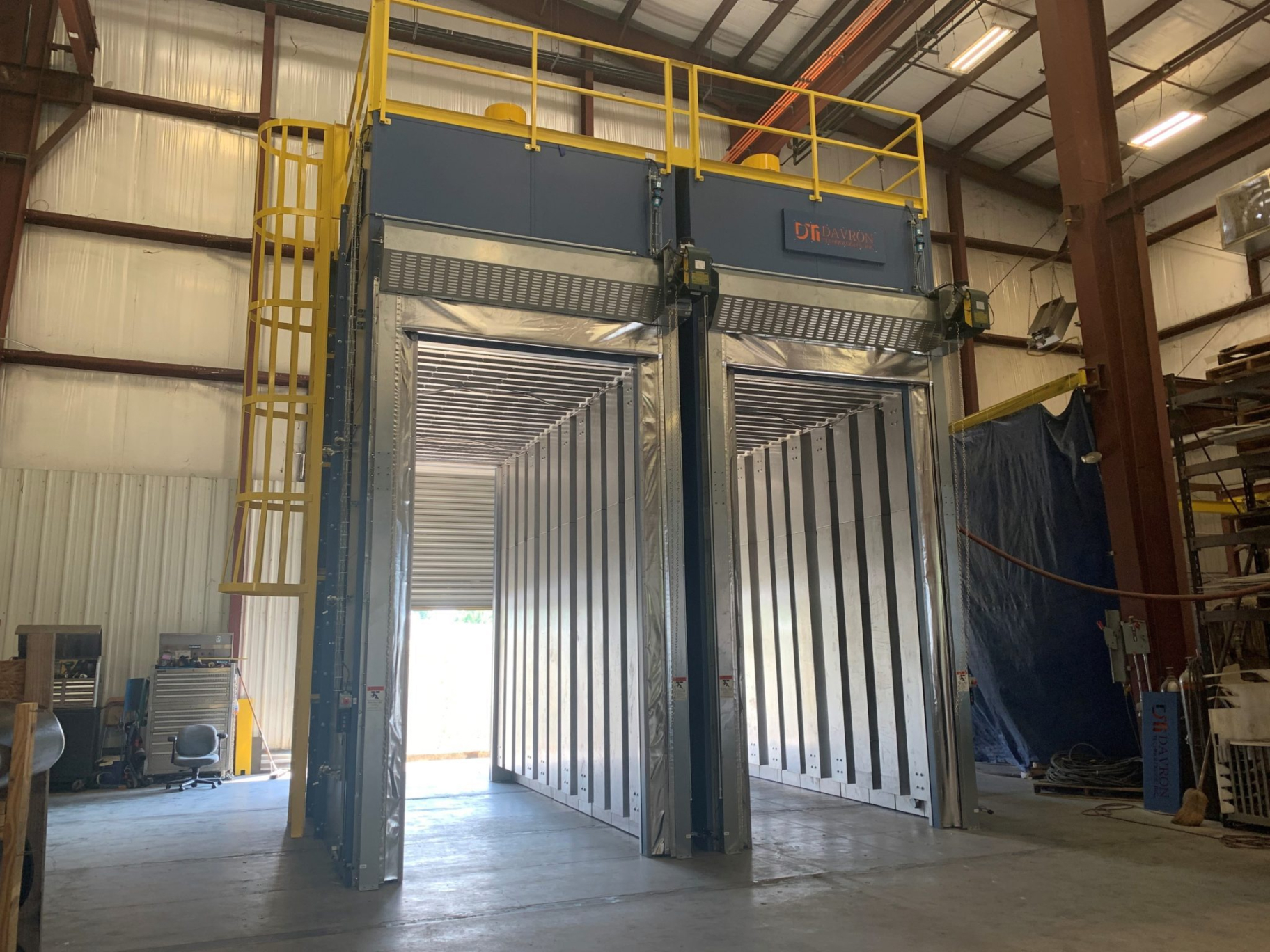 Interior view of the DTI-1478 Annealing Batch Oven, showing the large, open space inside the oven with vertically-oriented metal panels lining the walls. The entrance is wide, with tall, sliding doors fully opened to reveal the spacious interior designed for processing large amounts of extruded HDPE tubes and molded domes. A yellow safety ladder is visible on the left side, leading up to the top platform, which is surrounded by yellow railings. The industrial setting is evident, with tools and equipment visible in the background.