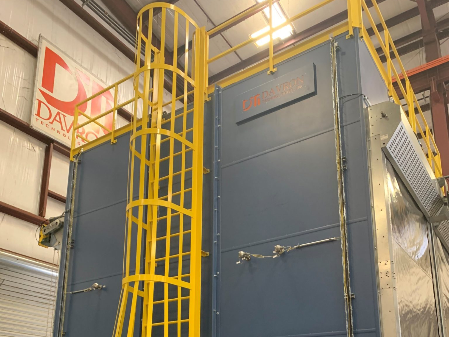 Close-up view of the DTI-1478 Annealing Batch Oven, focusing on the side and top sections. The exterior walls of the oven are highlighted, along with a yellow safety ladder that leads up to the roof, which is protected by yellow railings. A metal sign displaying the 'Davron Technologies Inc.' logo is mounted on the side of the oven. The industrial environment is visible in the background, including a portion of the facility’s interior structure.